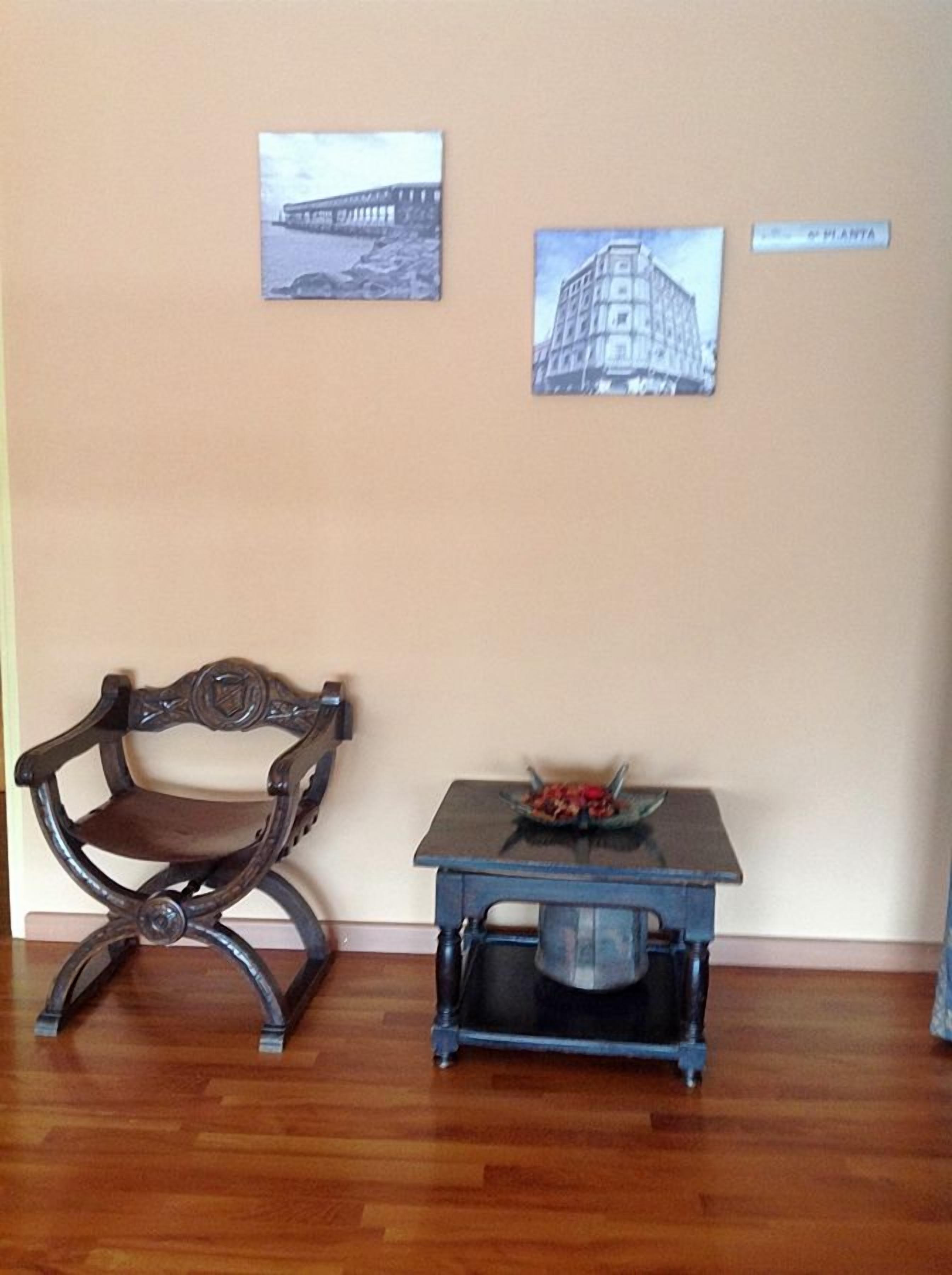 A room with a wooden floor, a dark wooden chair, a small table with a bowl of fruit, and two framed pictures on the wall.