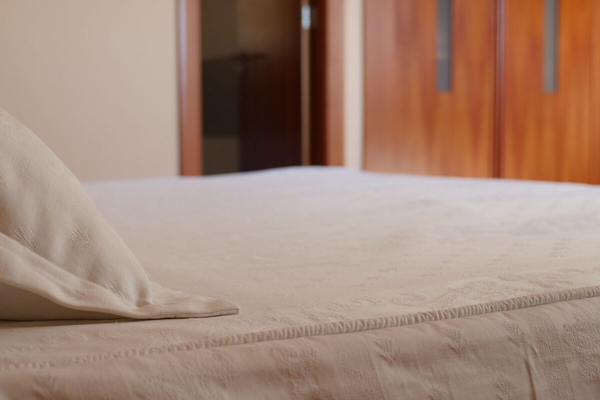 Una cama con un almohadón blanco y una manta texturizada en una habitación con puertas de madera y una pared lisa.