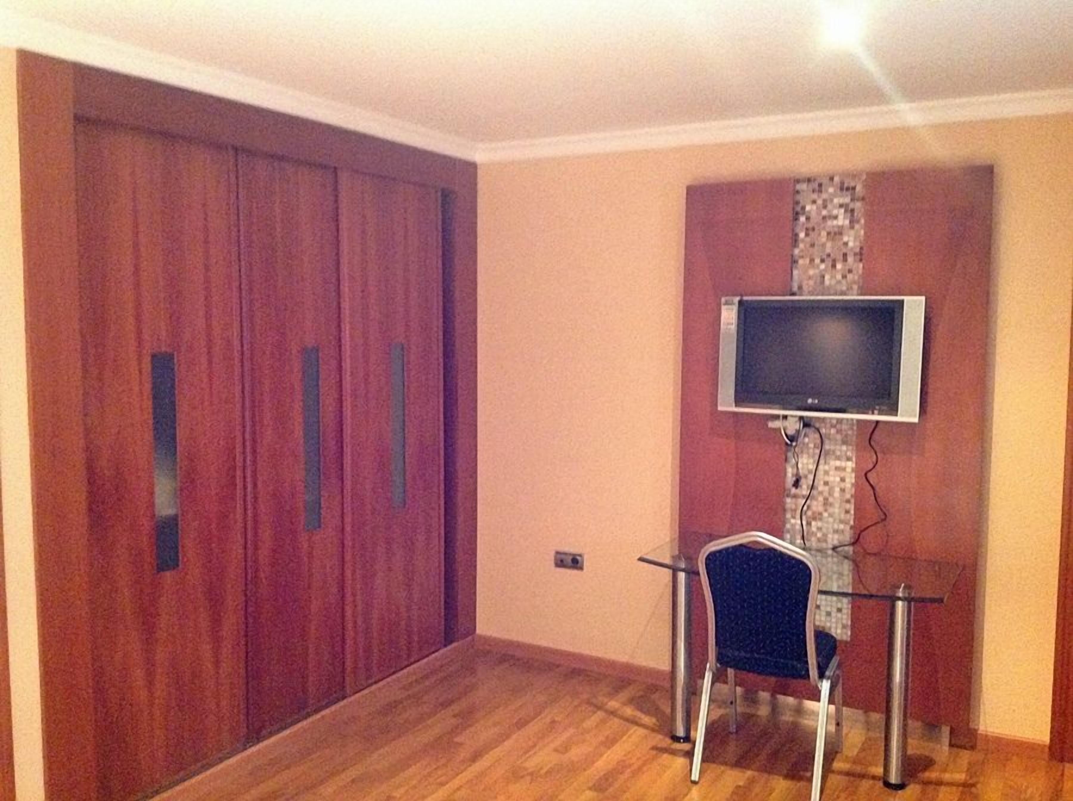 Una habitación con un armario de madera, un televisor montado en la pared y una mesa de vidrio con una silla.