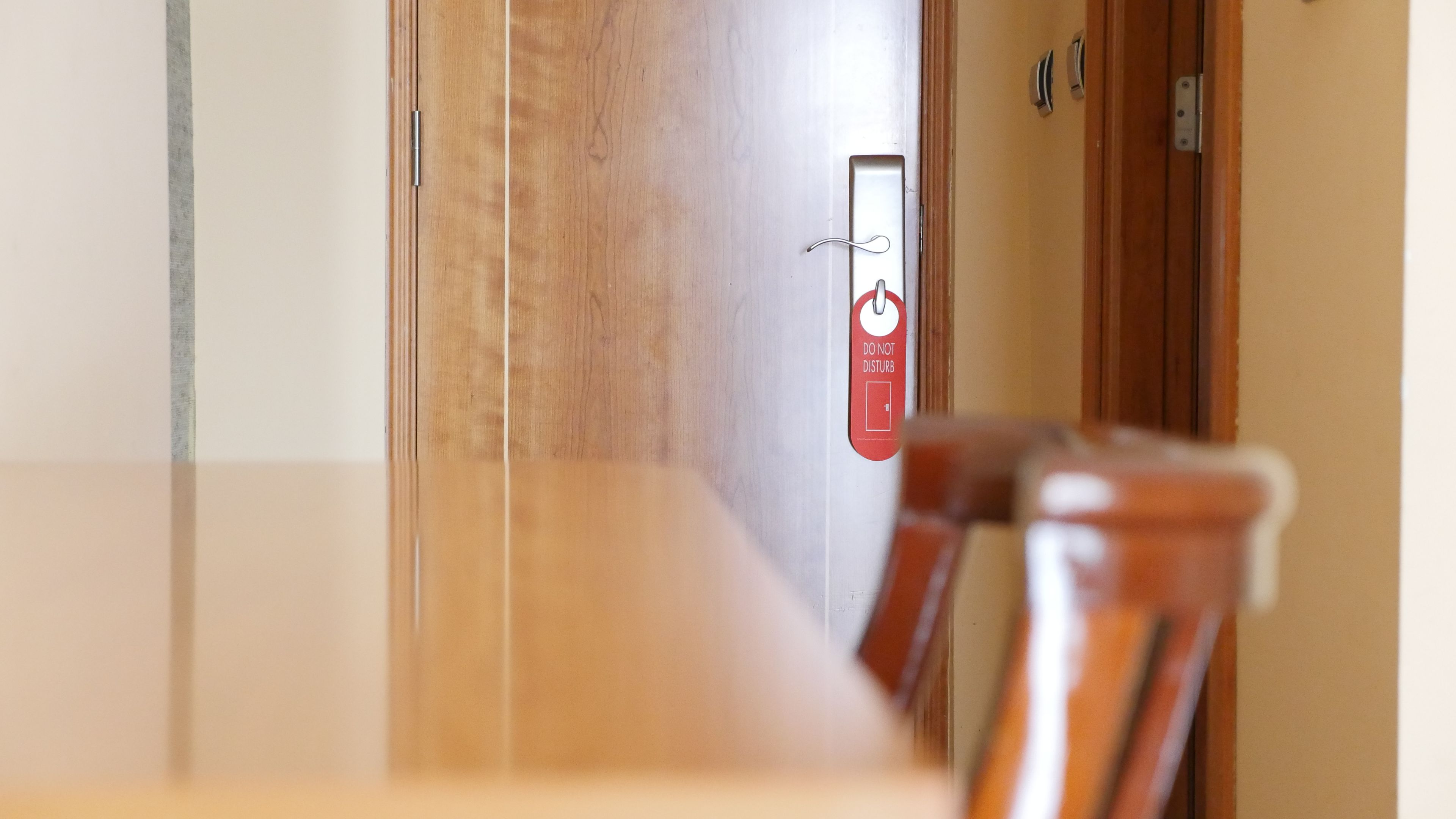 Habitación de hotel con una puerta de madera, un cartel rojo con un símbolo y texto, y una silla en primer plano.