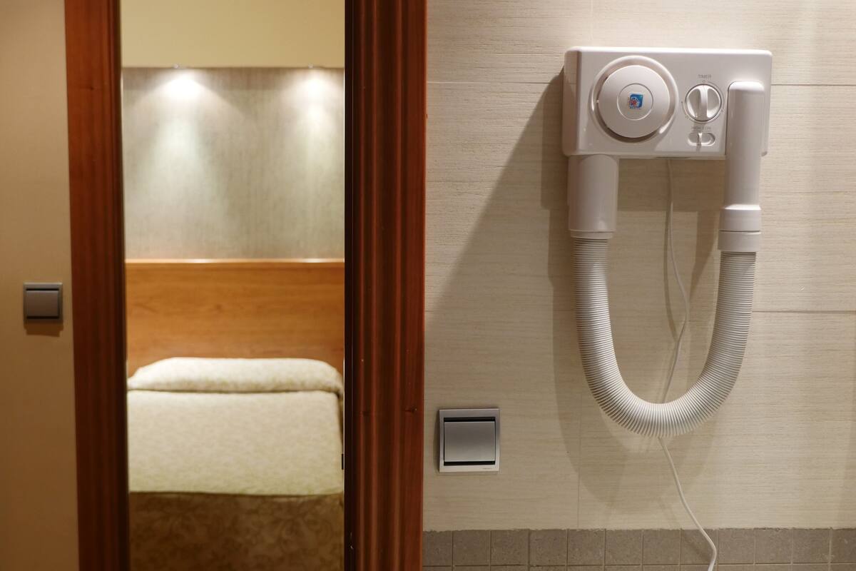 Ein Hotelzimmer mit Holztür, einem Bett mit weißer Matratze, einem an der Wand befestigten Heizkörper und einer Wandklimaanlage.