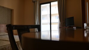 A hotel room with a wooden dining table, a chair, a television, and a view of a building through the window. - Rusadir (Melilla)