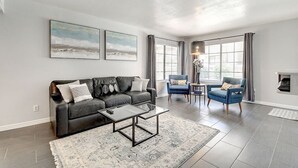 A modern living room with a black leather sofa, a coffee table, and a blue armchair. There are two large windows with curtains, a fireplace, and a rug on the floor.