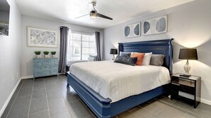 A bedroom with a large bed, a ceiling fan, a nightstand with a lamp, a dresser, and a window with curtains.