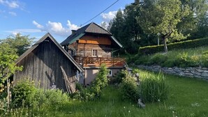 Een rustiek houten huis met schuin dak en een stenen muur, omgeven door weelderige begroeiing en een heldere blauwe lucht.