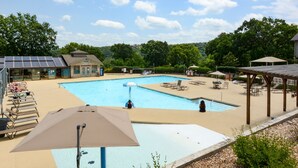 Ein Schwimmbecken mit Liegestühlen und Sonnenschirmen, umgeben von Bäumen und einem Gebäude mit Solaranlage.