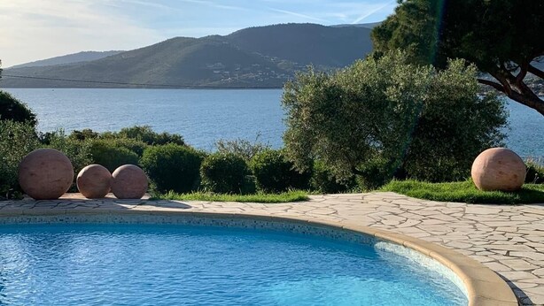Una piscina con borde de piedra, rodeada de follaje y vista a un lago y montañas.