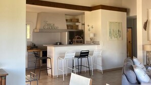 Un salón espacioso con una mesa de comedor de madera, sillas blancas y un sofá gris. Se ve una cocina con armarios blancos y techo de madera al fondo.