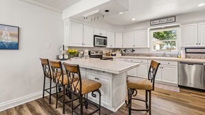 A modern kitchen with white cabinetry, a central island with bar stools, a stainless steel dishwasher, and a sign that reads "Live Love Lake". - Spotless 2-Bedroom Cottage, Highly Recommended (Mabank)