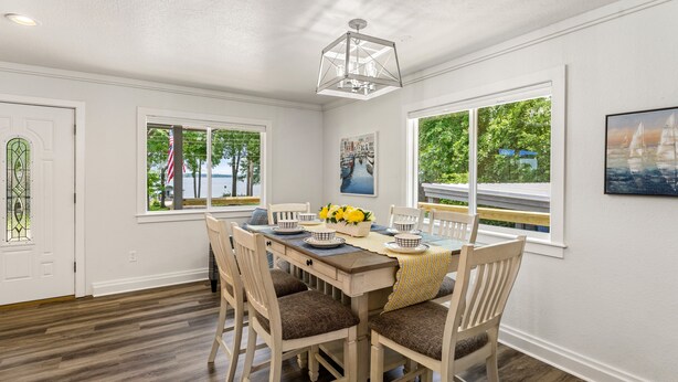 A dining room with a wooden table set for four, surrounded by white chairs with brown cushions. There are two windows with views of greenery and a sailboat. - Spotless 2-Bedroom Cottage, Highly Recommended (Mabank)