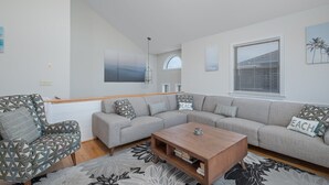 Un salon moderne avec un canapé d’angle, un fauteuil à motifs, une table basse en bois et une grande fenêtre avec des stores.