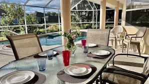 A glass-top table with two plates, two glasses, and a vase with flowers, set in a covered outdoor area with a pool in the background.