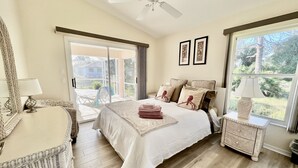A bedroom with a bed, a bedside table, a chair, a ceiling fan, and large windows.