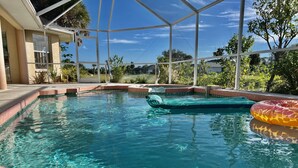 A clear swimming pool with a green inflatable float, surrounded by a covered patio area with a view of greenery and a building in the background.