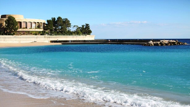 A beach with clear turquoise water, a sandy shore, and a building with arched windows in the background.