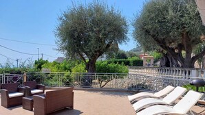An outdoor seating area with wicker furniture and white lounge chairs, surrounded by trees and a stone wall.