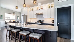 Een moderne keuken met een groot eiland, barstoelen, hanglampen en een bordje voor de voorraadkast.