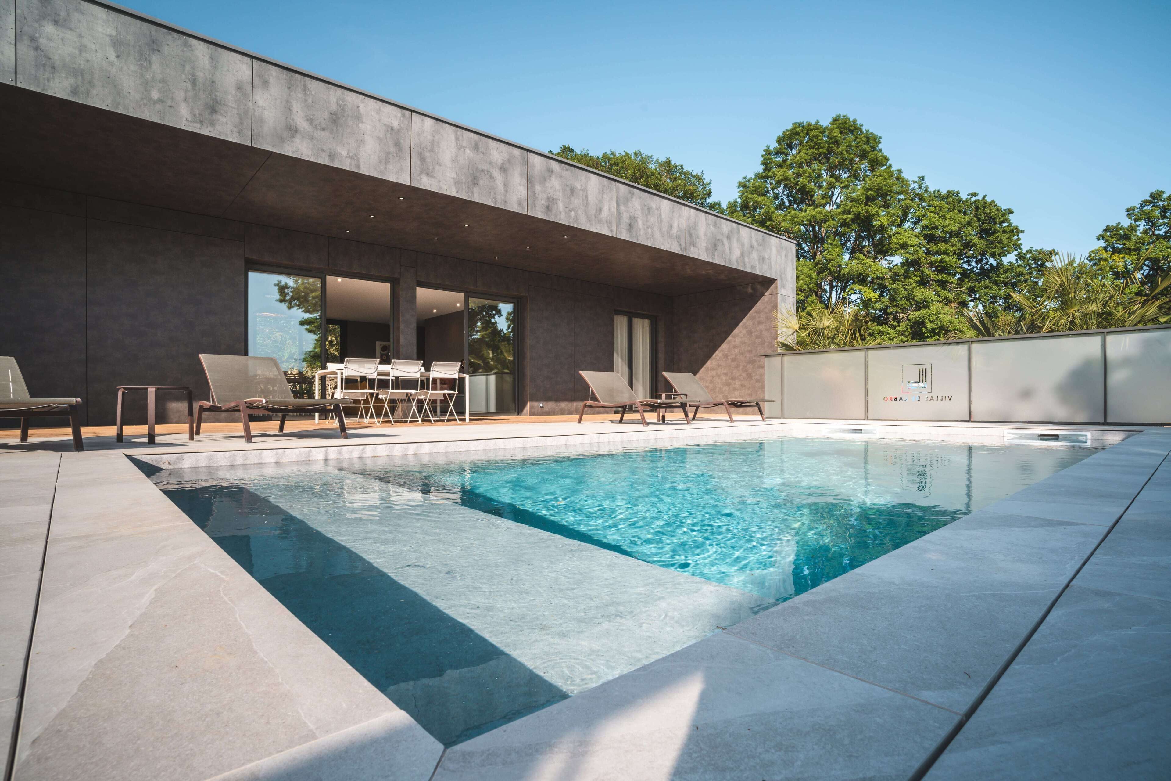 Un área de piscina moderna al aire libre con un suelo de concreto, sillas de descanso y una mesa de comedor con sillas.