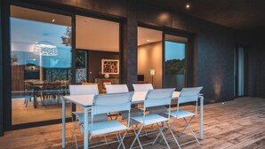 Un espace de restauration extérieur moderne, doté d’une terrasse en bois, de chaises en métal blanc et d’une porte en verre donnant accès à un espace intérieur comprenant une cuisine et une table à manger.