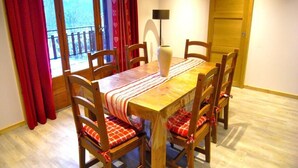 Un coin repas avec une table et des chaises en bois, un vase posé sur la table et une vue par une fenêtre ornée de rideaux rouges.