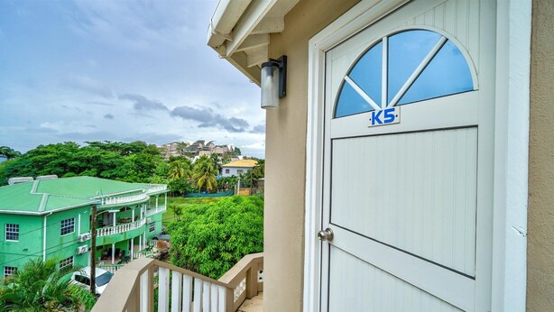 A white door with a blue 'K5' sign, a green building with a balcony, and lush greenery in the background. - Quaint one bedroom unit steps away from beach access (Saint George)