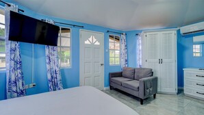 A bedroom with a bed, a grey sofa, a wardrobe, and a TV mounted on the wall. - Quaint one bedroom unit steps away from beach access (Saint George)