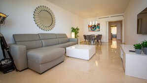 A modern living room with a grey sofa, a white coffee table, a large mirror, and a dining area with chairs and a table.
