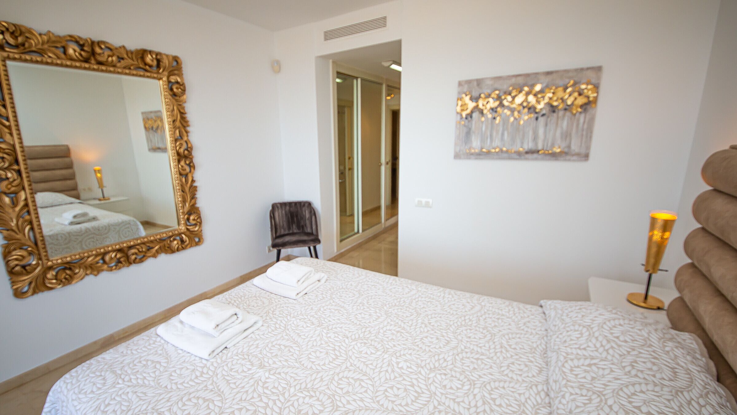 A bedroom with a large ornate mirror, a bed with white linens, a small chair, and a bedside table with a lamp.
