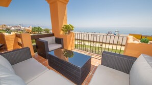 A balcony with wicker furniture, a glass table, and a view of the sea and beach.
