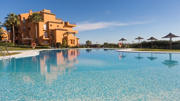 A swimming pool with clear blue water, surrounded by a building with balconies and palm trees.