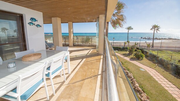 Un balcón con vista a la playa y al océano, una mesa con sillas y una ventana con peces decorativos.