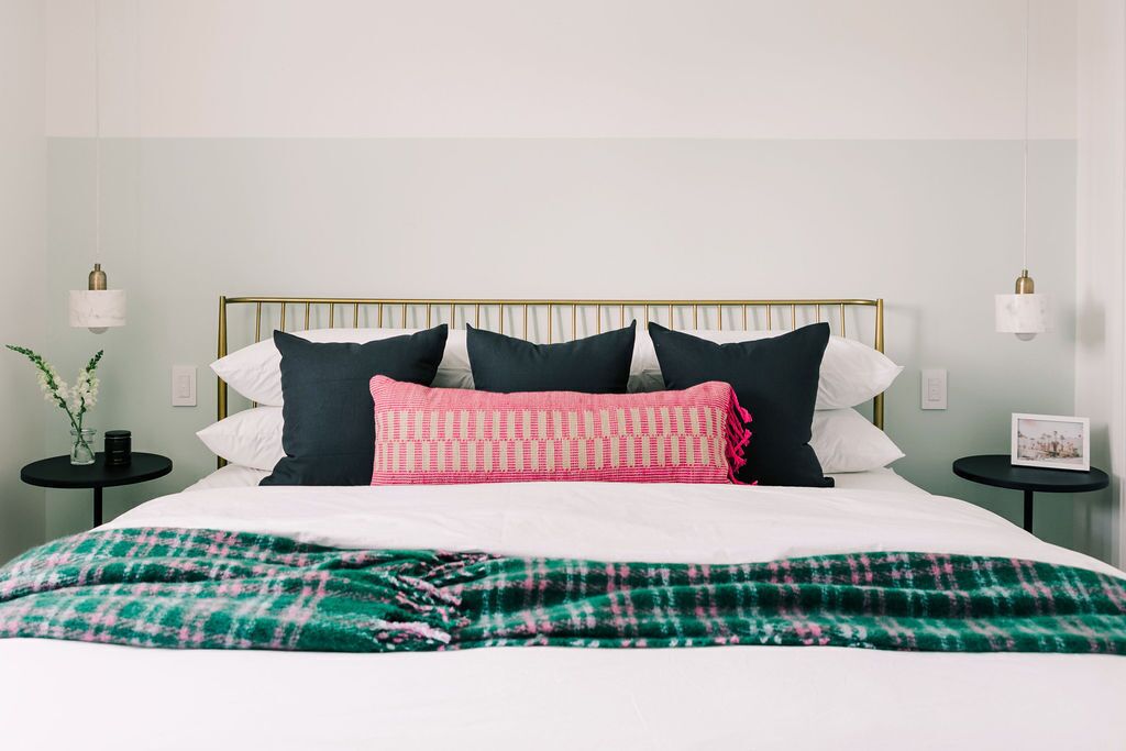 Een net opgemaakt bed met een groen-wit geruit dekbed, een gouden hoofdbord en meerdere kussens in tinten wit, zwart en roze.