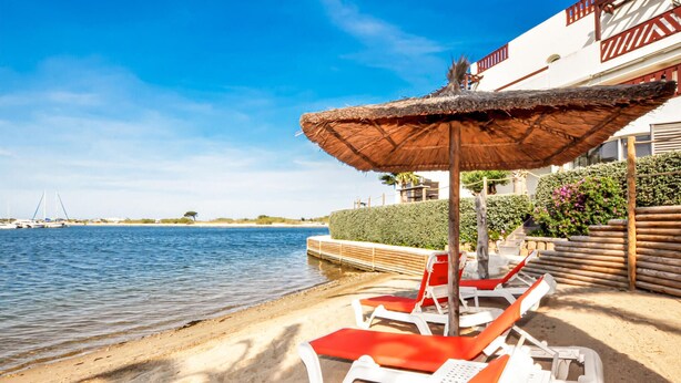 Een loungeruimte aan het strand met ligbedden en een rieten parasol, met uitzicht op een kalme zee en een wit gebouw met rode accenten.