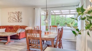Ein gemütliches Wohnzimmer mit einem roten Ledersofa, einem Holzesstisch und Stühlen. Ein großes Fenster bietet Blick ins Grüne.