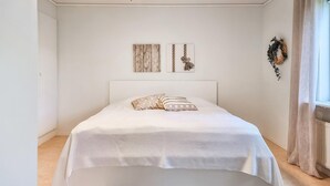 Ein Schlafzimmer mit einem Bett, einem Kopfteil, einer Wand mit zwei gerahmten Bildern und einem Fenster mit Vorhängen.