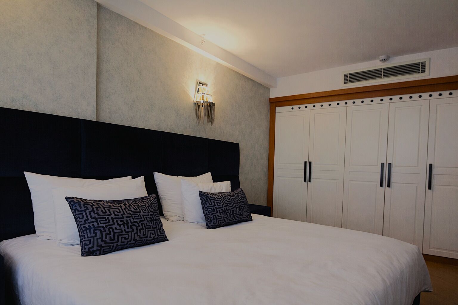 A bedroom with a large bed, a headboard, a wardrobe, and a wall-mounted light fixture.
