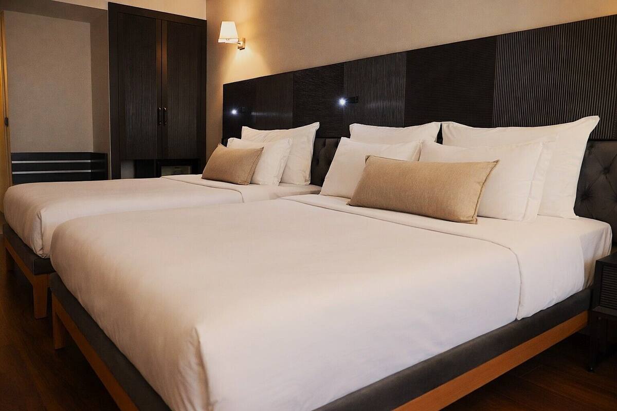 A double bed with white and beige bedding, a dark headboard, and a wall-mounted lamp.