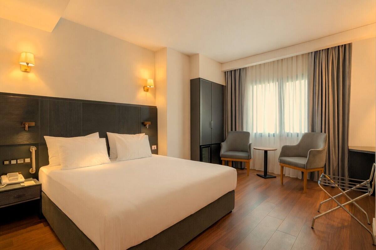 A hotel room with a large bed, two armchairs, a small table, a wardrobe, and a desk with a telephone.