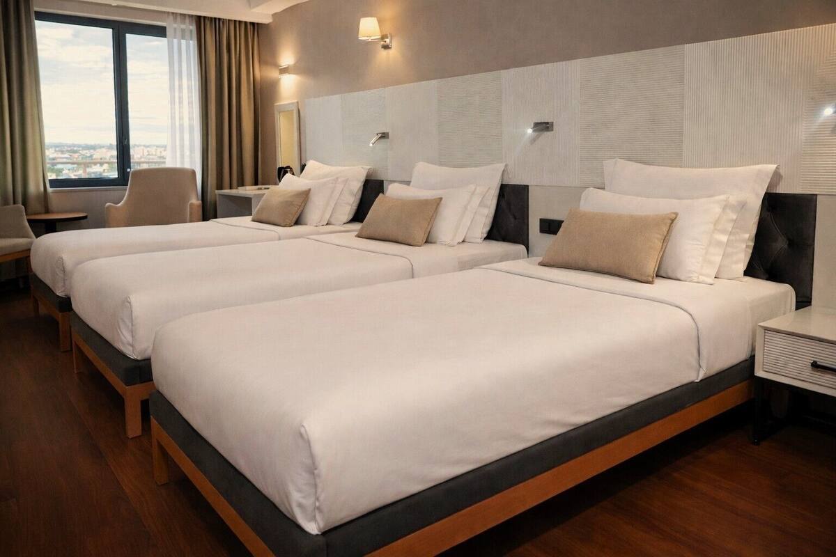A hotel room with two beds, a desk, a chair, and a large window offering a city view.