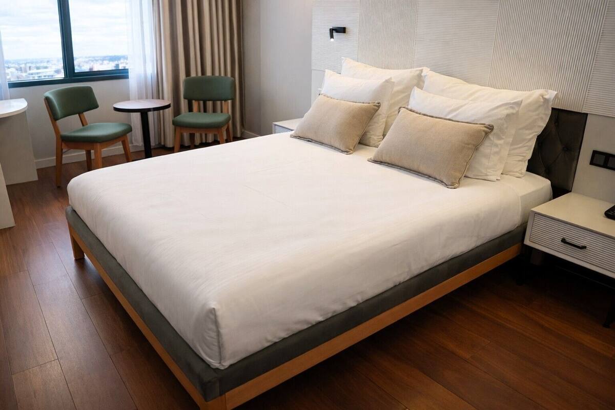 A modern hotel room with a large bed, a bedside table, a chair, and a view of the city through the window.