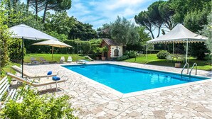 Une piscine entourée de chaises longues et de parasols, le tout bordé de verdure et adossé à un petit bâtiment en briques.