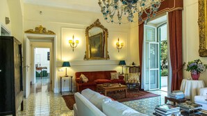 Un salon spacieux avec un canapé rouge, un fauteuil blanc et une table basse. On y trouve deux lampes fixées au mur, un grand miroir et un lustre. Le sol est recouvert de carreaux décoratifs et une grande fenêtre offre une vue sur l’extérieur.