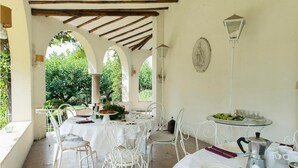 Un espace extérieur abrité, aménagé pour les repas, avec des tables et des chaises blanches, une cafetière et une vue sur la verdure à travers des arcades.