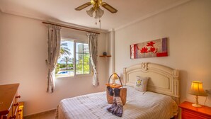 A bedroom with a bed, a ceiling fan, a window with curtains, a painting, and a nightstand with a lamp. - Cozy apartment in La Mata, Torrevieja (La Mata, Torrevieja)