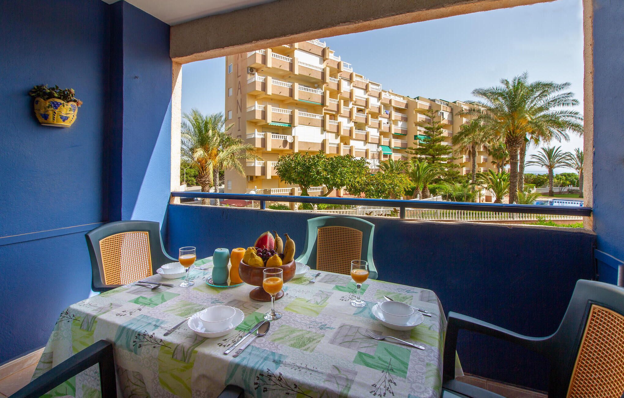 A dining area with a table set for two, a view of a building with balconies, and a palm tree in the background.