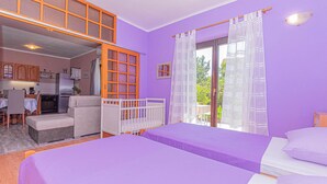 A room with a purple wall, a white bed, a white crib, a kitchen area with a dining table, and a window with curtains. - Awesome apartment in Raslina (Raslina)