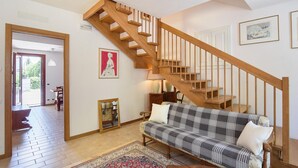 Een woonkamer met een houten trap, een bank, een vloerkleed en kunstwerken in lijsten aan de muren.