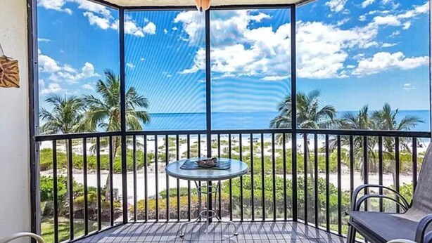 A balcony with a table and chairs, overlooking a beach with palm trees and the ocean. - Gulf Front Beach Life (Fort Myers Beach)