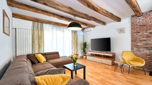 Un salon moderne avec des poutres apparentes, un téléviseur à écran plat, un ensemble de canapés, un fauteuil jaune et un mur décoratif en briques.