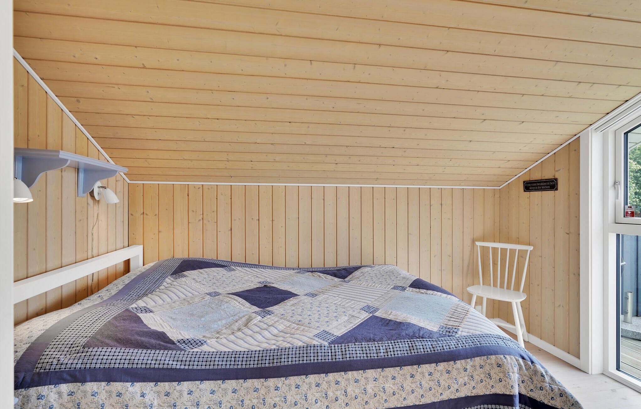 A bedroom with a wooden ceiling, a bed with a patterned quilt, a white chair, and a window with a view of greenery.